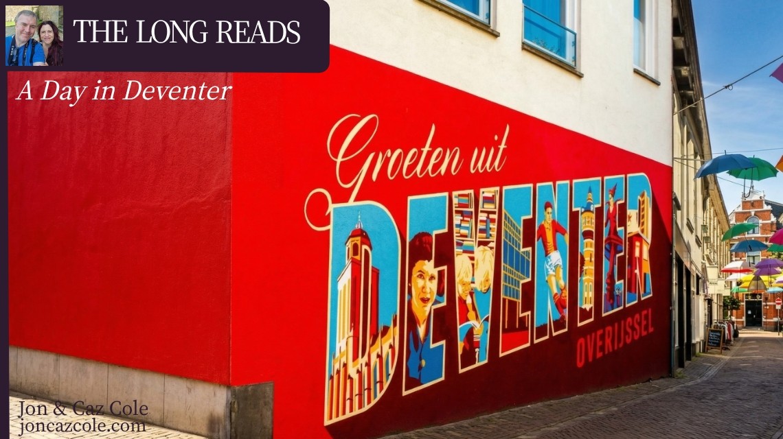 A vibrant red street mural in Deventer, Netherlands, featuring the town's name in illustrated letters, with colorful umbrellas hanging over a medieval cobblestone alleyway