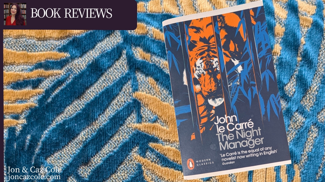 A landscape book review banner for the blog Jon & Caz Cole, featuring the Penguin Modern Classics edition paperback of John le Carré’s spy thriller The Night Manager. The book cover, illustrated with a striking orange tiger behind dark blue bamboo stalks, is set against a vibrant background of textured teal and gold fabric. A dark plum "Book Reviews" badge sits in the top left corner.